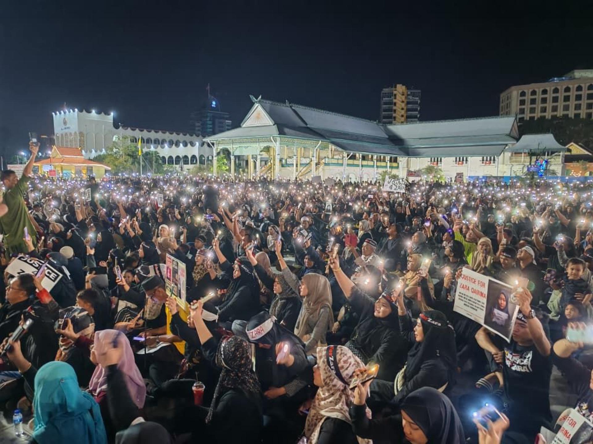 民众席地而坐响应主持人的呼吁，开启手机的手电筒功能，一起高喊“我们疼爱扎拉”。