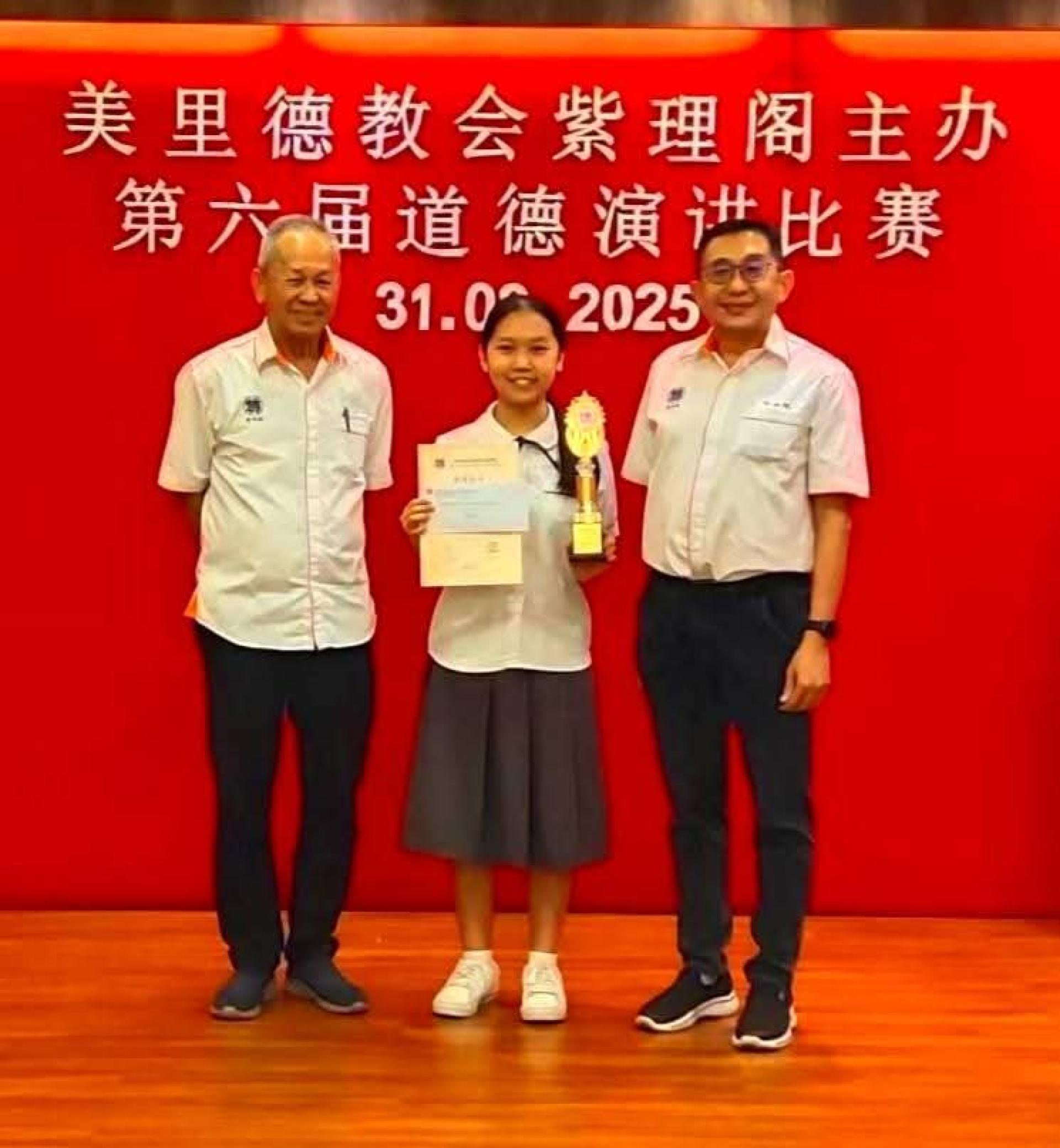 美里德教会紫理阁第六届道德演讲比赛，培中生陈家瑾获得初中组冠军。左为署理阁长何洪坤，右为教育主任兼筹备会主席许达伟。