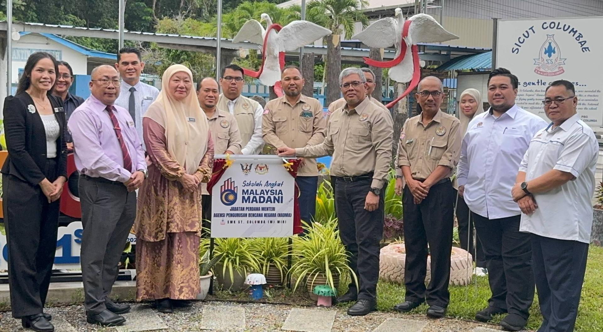嘉宾与学校领导层在昌明领养学校校牌前合照,象征合作关系,共同推动教育发展。前排右4为NADMA副总监(灾后管理组)哈芝胡赛因,前排左1为圣哥伦巴中学行政副校长夏碧燕,前排左2为圣哥伦巴中学校长金本马桑,前排左3为美里县教育局局长玛丽安。