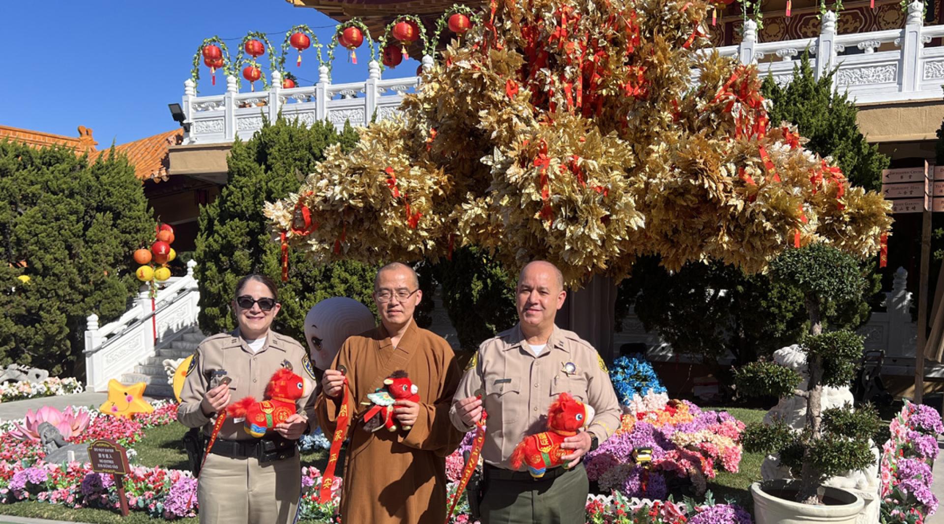 美国佛光山西来寺住持慧东法师（中）在成佛大道旁的诸事圆满祈愿树前，同负责春节期间交通与治安的当地警方人士合影。（图：中新社）