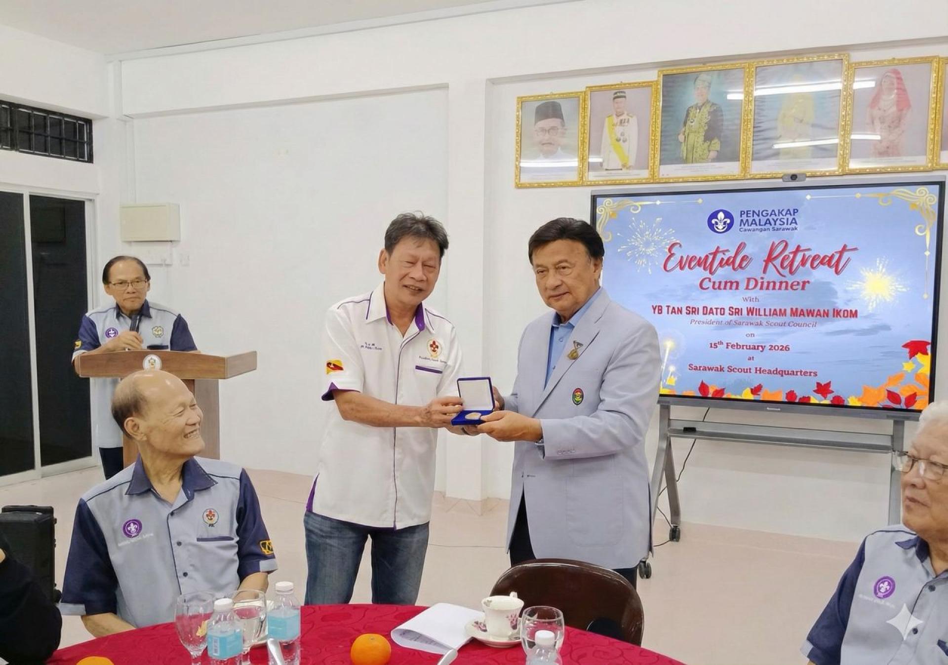 古晋市区童军理事会会长李江勝赠送纪念品徽章予丹斯里拿督斯里威廉玛旺，顾问许毓德（坐者）在旁见证。