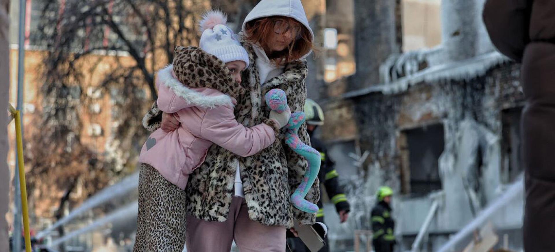 一名母亲带着女儿走过基辅一栋在夜间袭击中严重受损的工业建筑，消防员仍在向这栋建筑的残骸喷水。