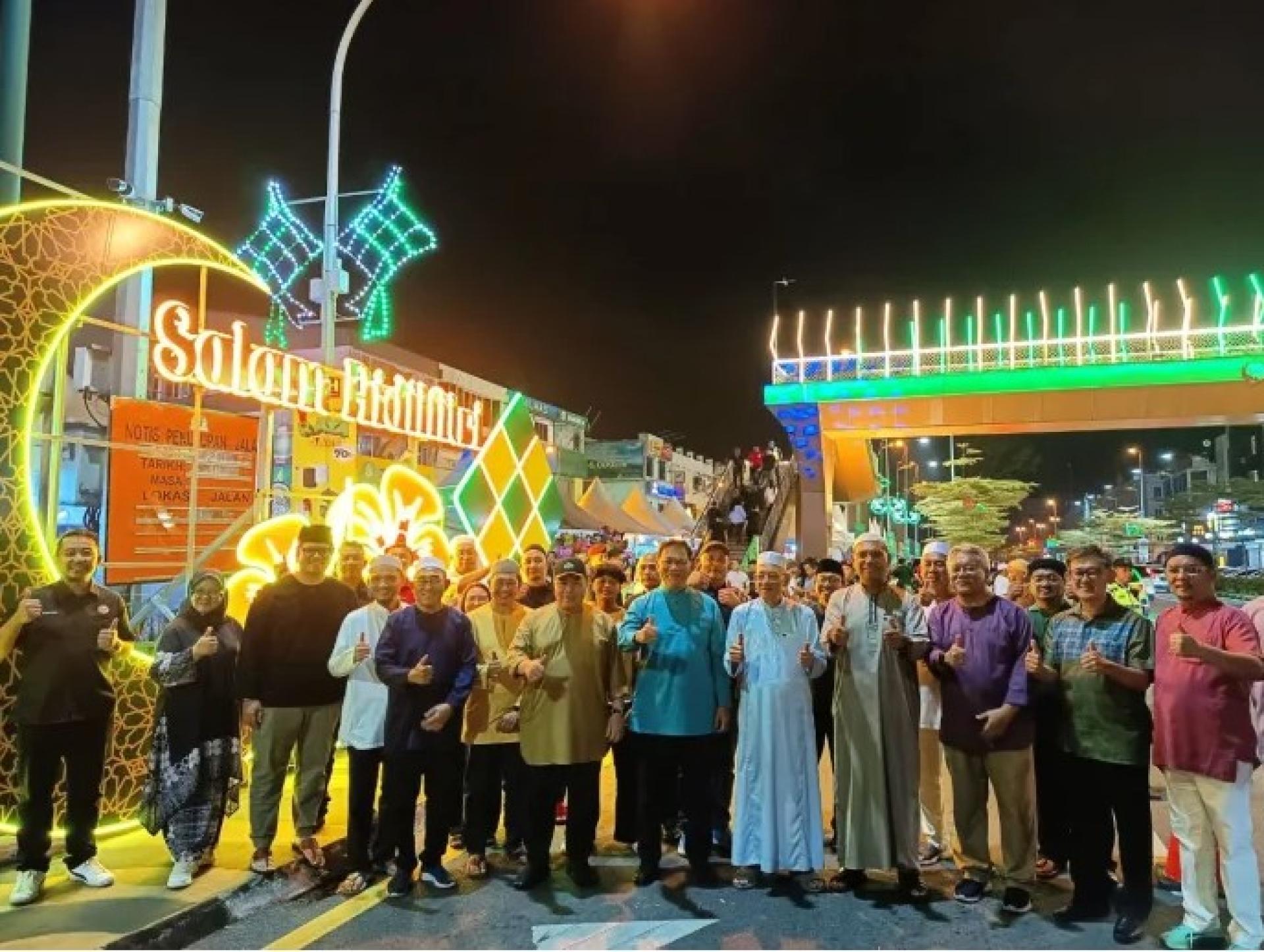 前排左起第七位起为希尔米、阿都拉曼及依布拉欣，与众人在砂督开斋节市集大型字样前合影，背景为亮灯的砂督行人天桥及装饰灯。（2026年3月9日）